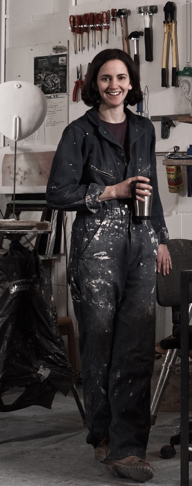 A female sculptor wearing paint-spattered overalls in her studio